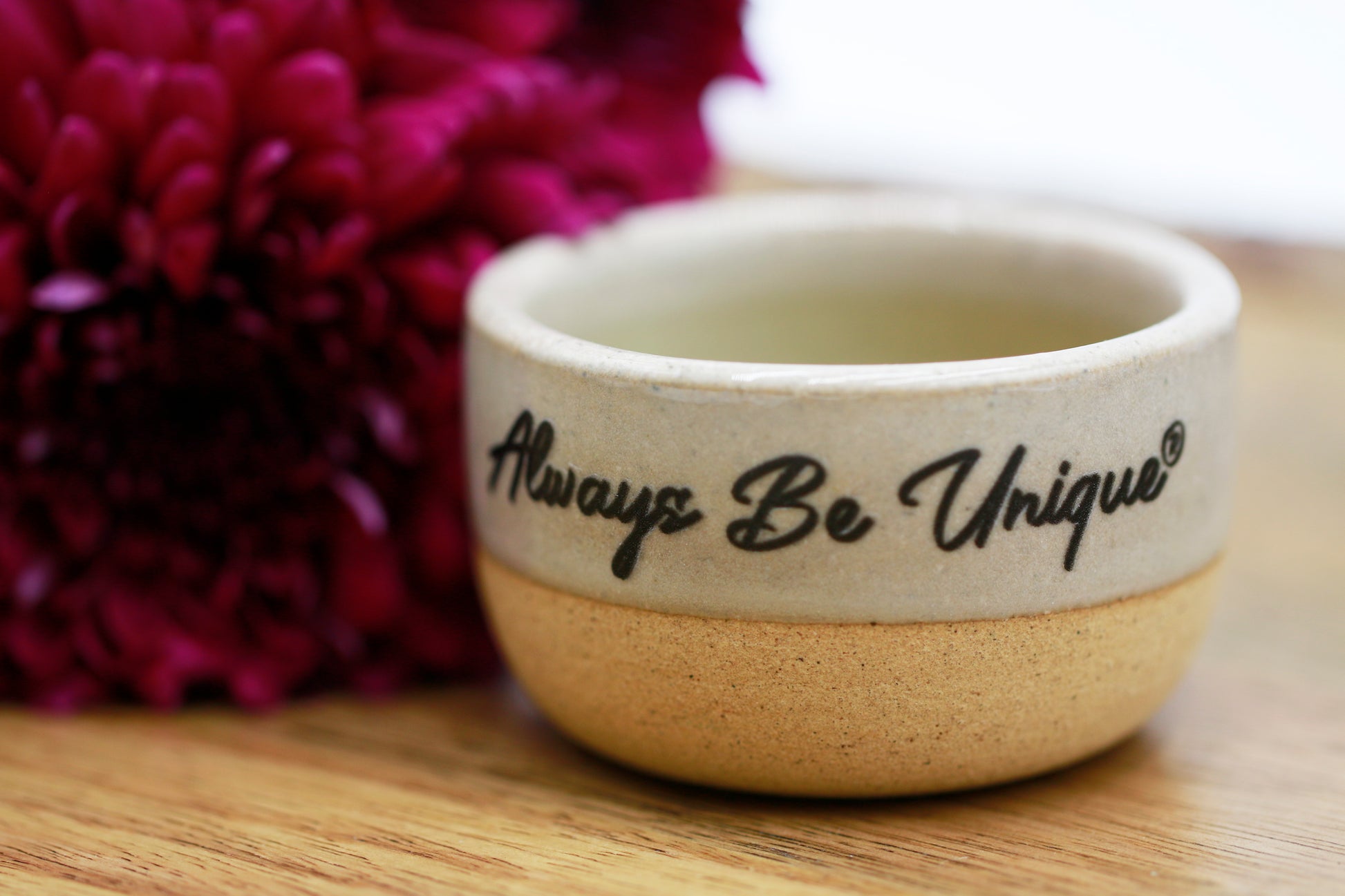 Small ceramic bowl with 'Always Be Unique' text on a wooden surface with flowers in the background